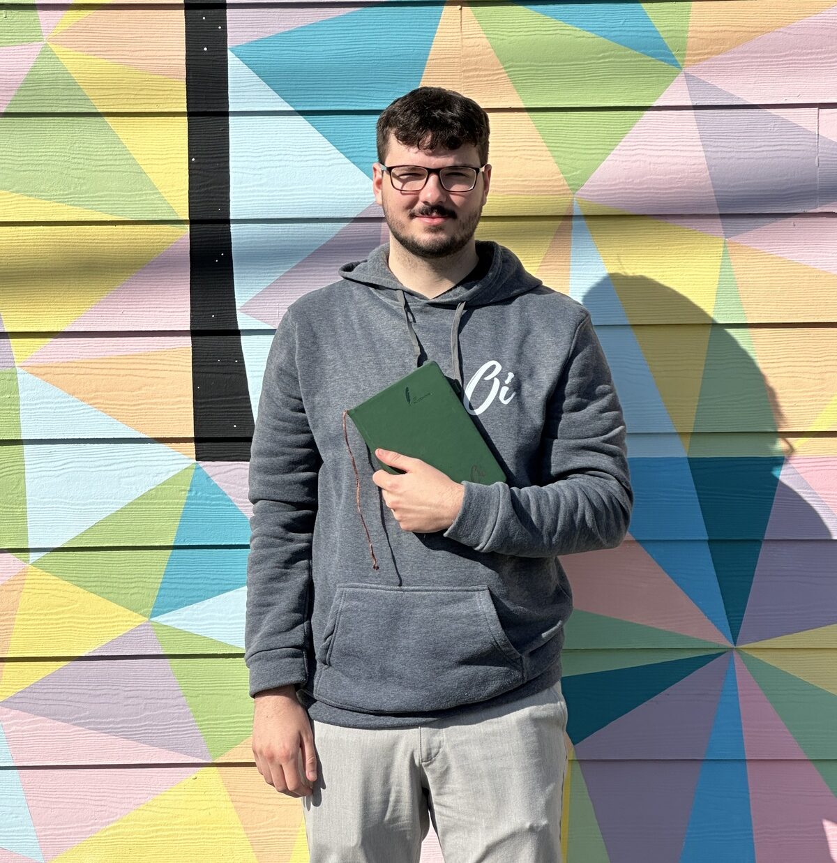 Our Team 7 Person with glasses and a beard stands holding a green book in front of a colorful geometric mural painted on horizontal planks.